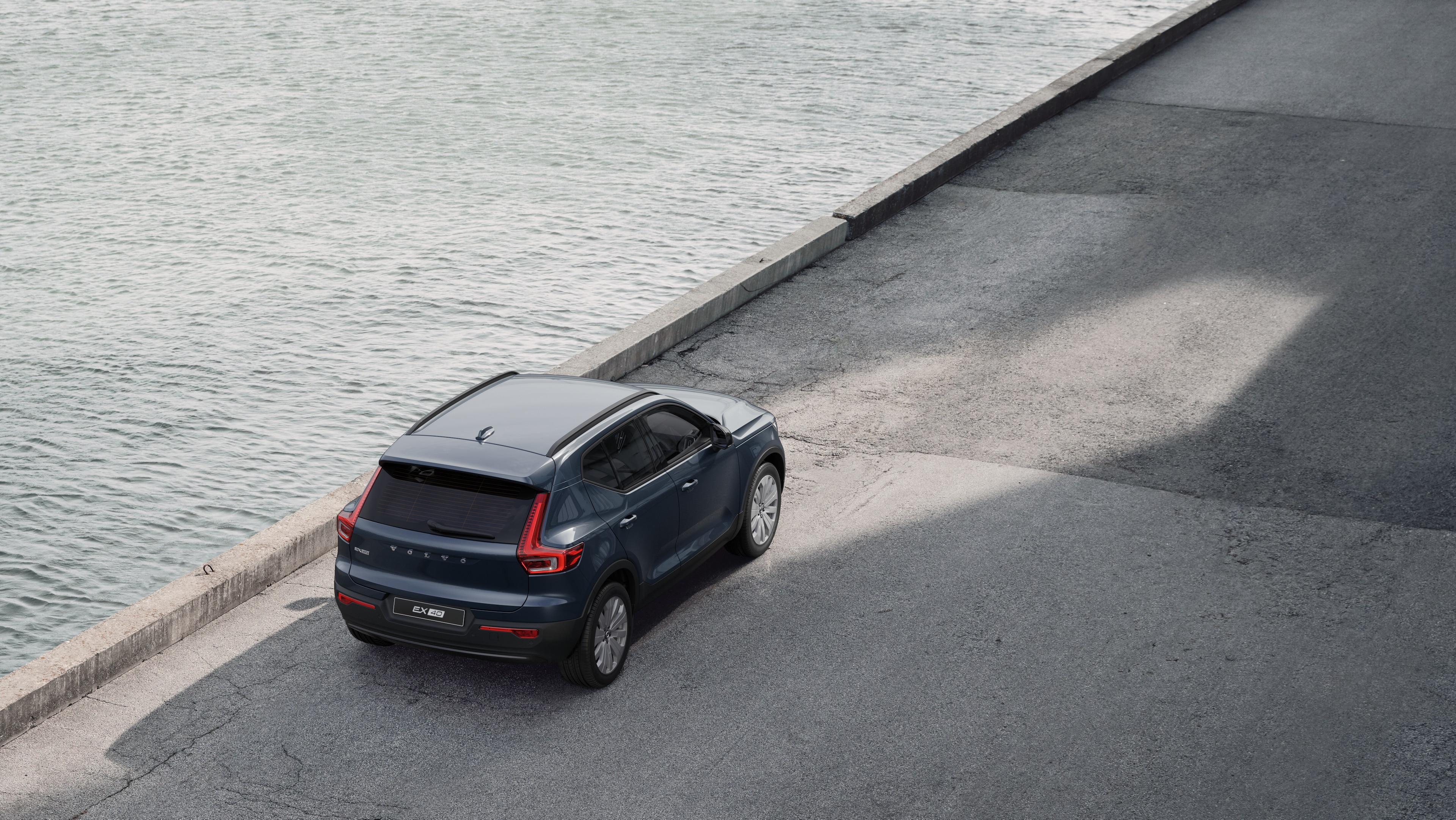 Volvo-bil set bagfra parkeret på en stille gade ved siden af vandet med en asfalteret gangbro og vand som baggrund.