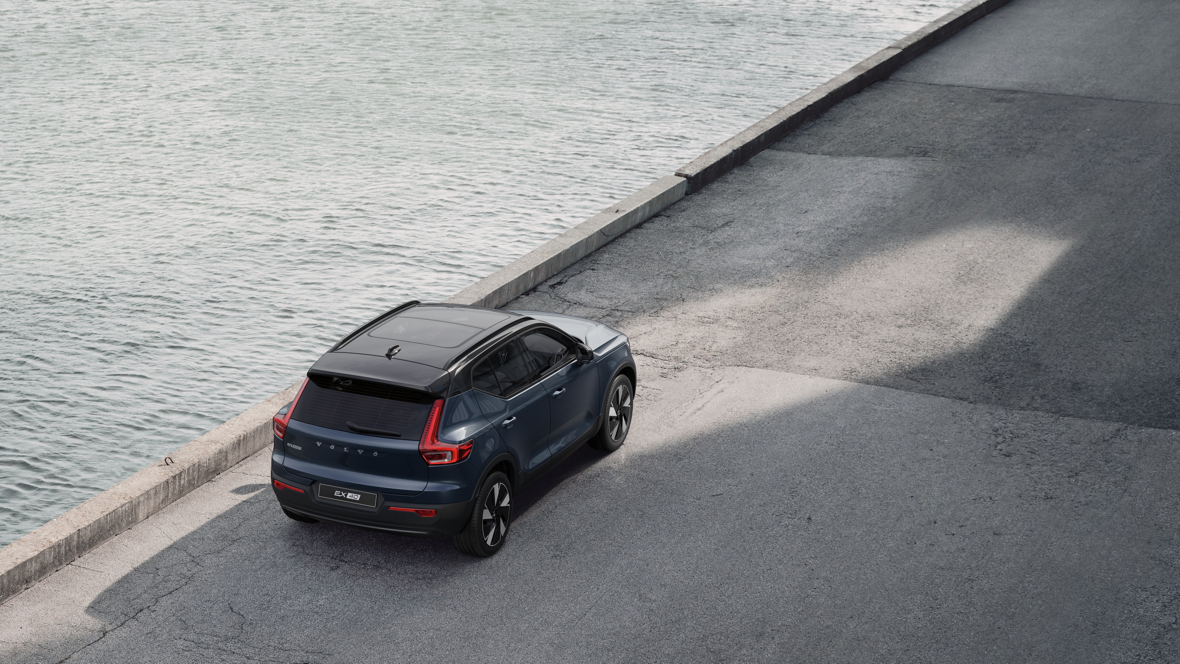 Volvo-bil set bagfra parkeret på en stille gade ved siden af vandet med en asfalteret gangbro og vand som baggrund.